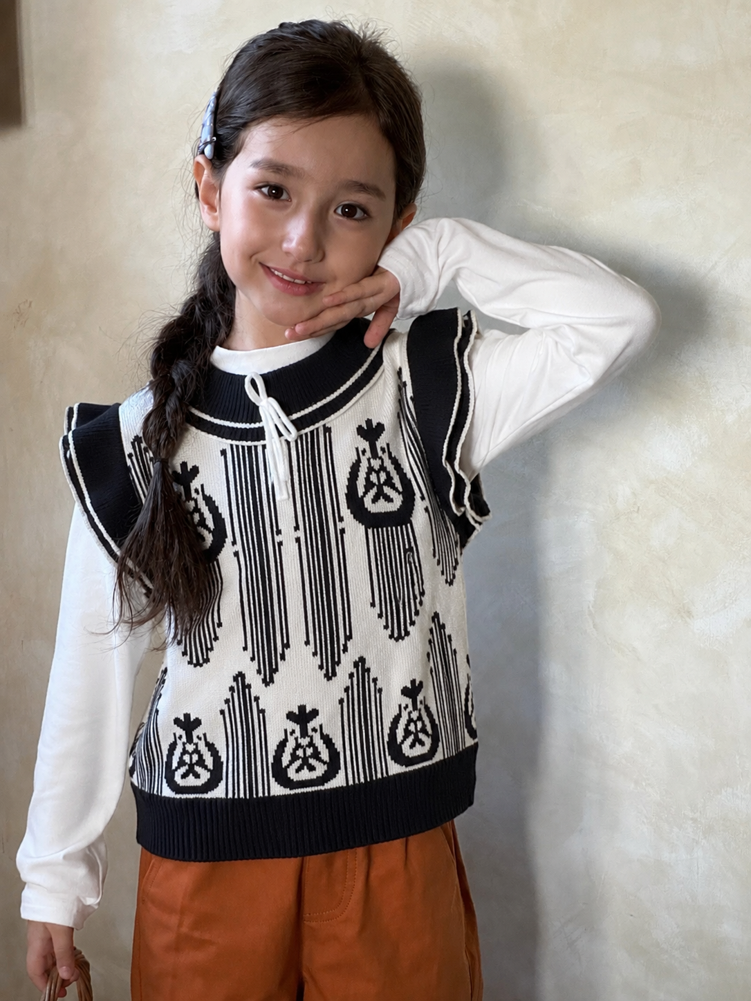 Girl with a long braid wearing the black and white ikat knit vest over a white shirt and rust pants indoors
