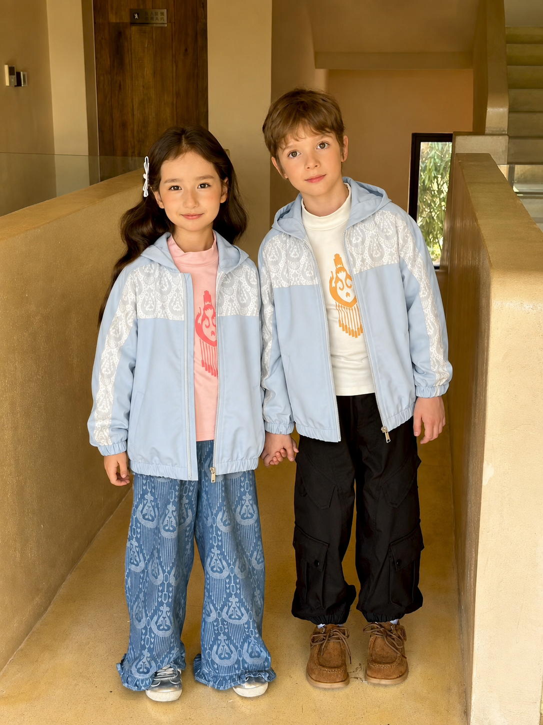 Two children, a girl and a boy, standing in a hallway wearing light blue hooded windbreakers with white ikat-patterned yokes.