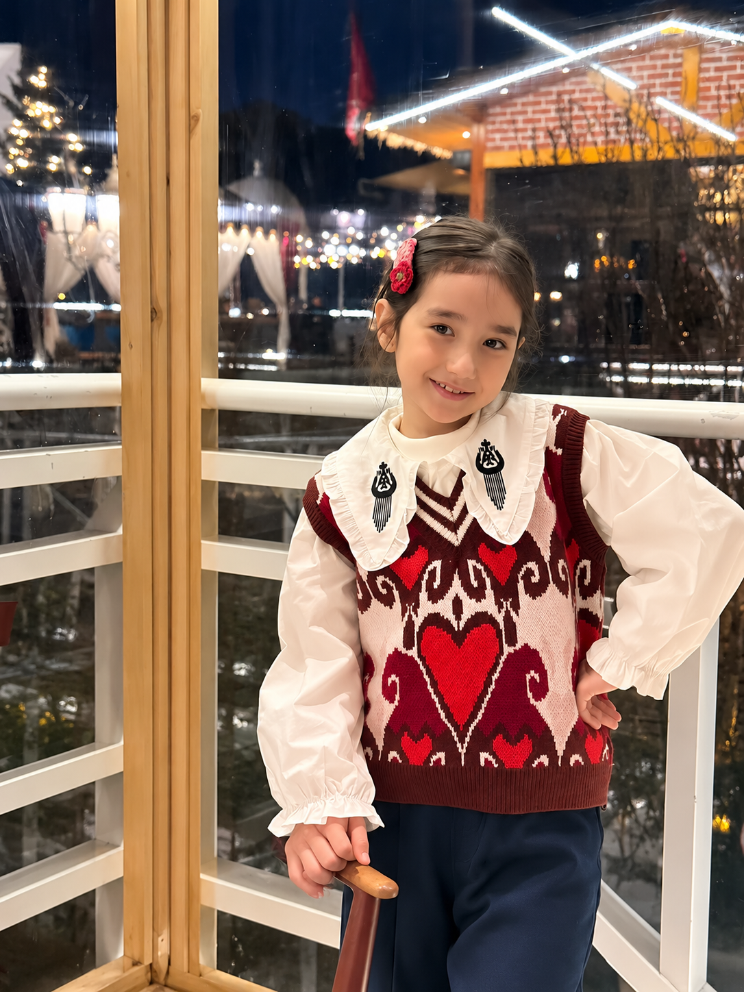 Young girl wearing the red atlas heart knit vest over a white blouse, standing on a terrace decorated with night lights