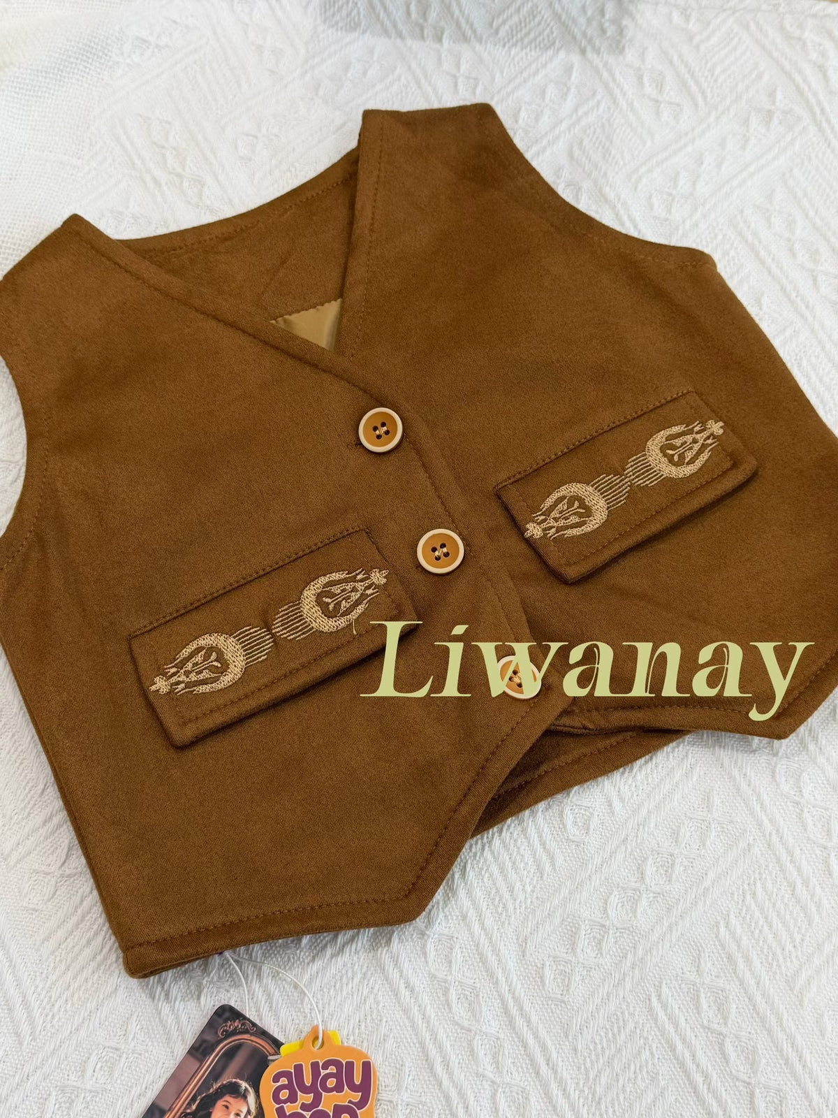 Brown suede-inspired vest with embroidered pockets and wooden buttons, laid flat on a white fabric background.