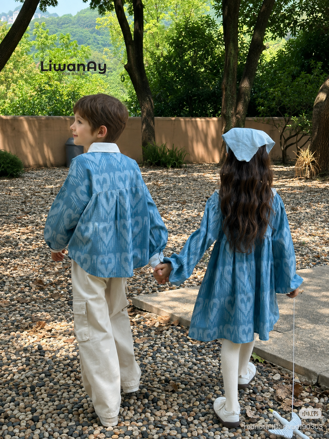 Back view of two children in matching blue ikat outfits walking hand in hand on a gravel path outdoors