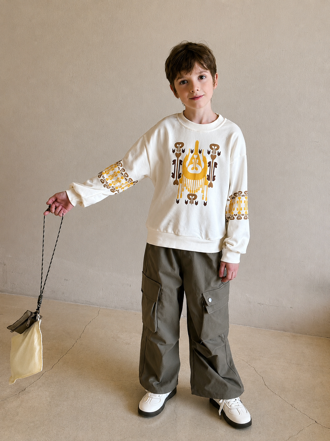 Boy wearing the cream atlas graphic sweatshirt with yellow ikat print, holding a small beige bag in a cafe