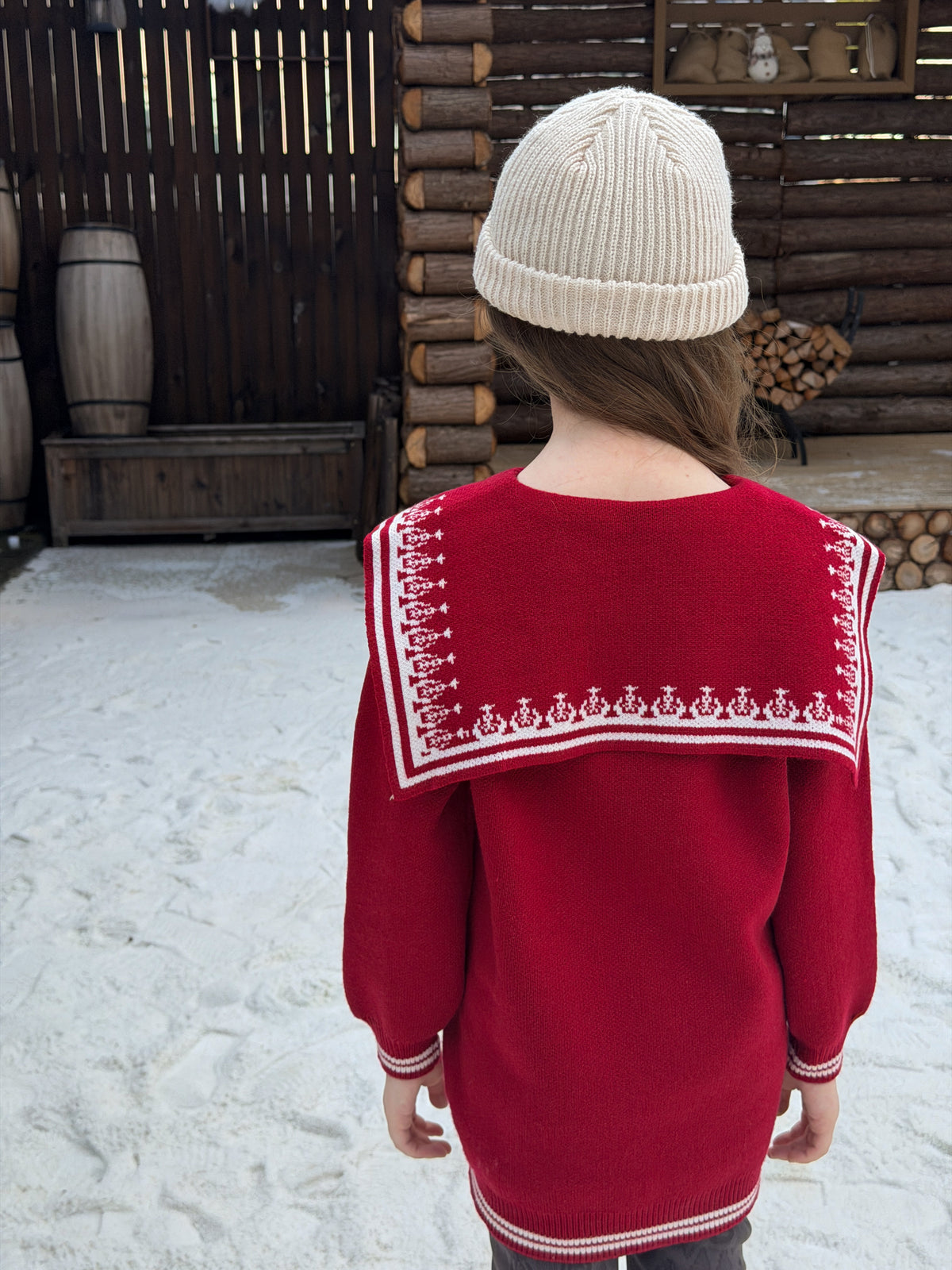 Back view of the red knit sweater dress showing the white embroidered yoke, worn with a cream beanie in a snowy cabin yard