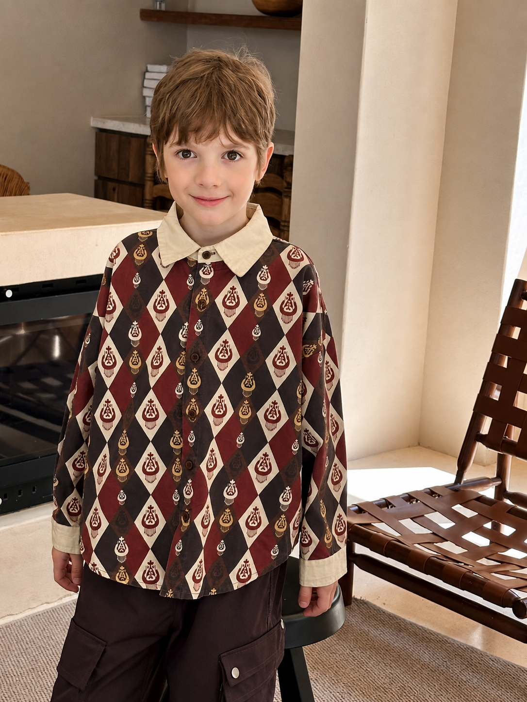 Boy standing by a fireplace in the burgundy diamond-ikat shirt with a cream collar and brown pants.