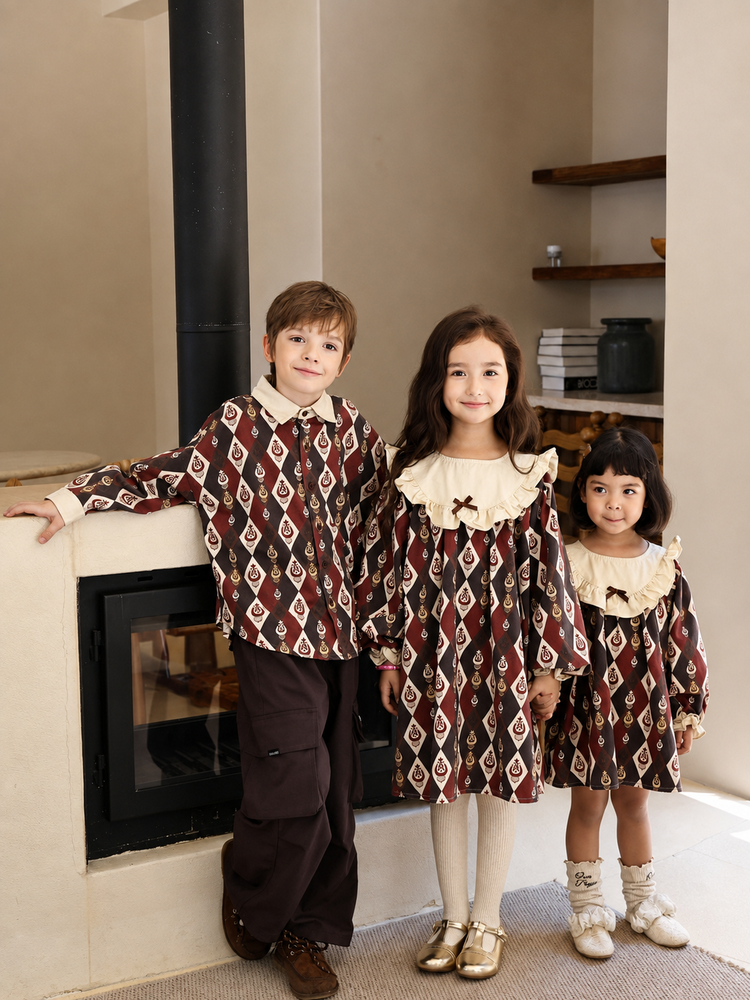 Three children in burgundy diamond-ikat outfits with cream ruffle collars, posing together beside a fireplace.