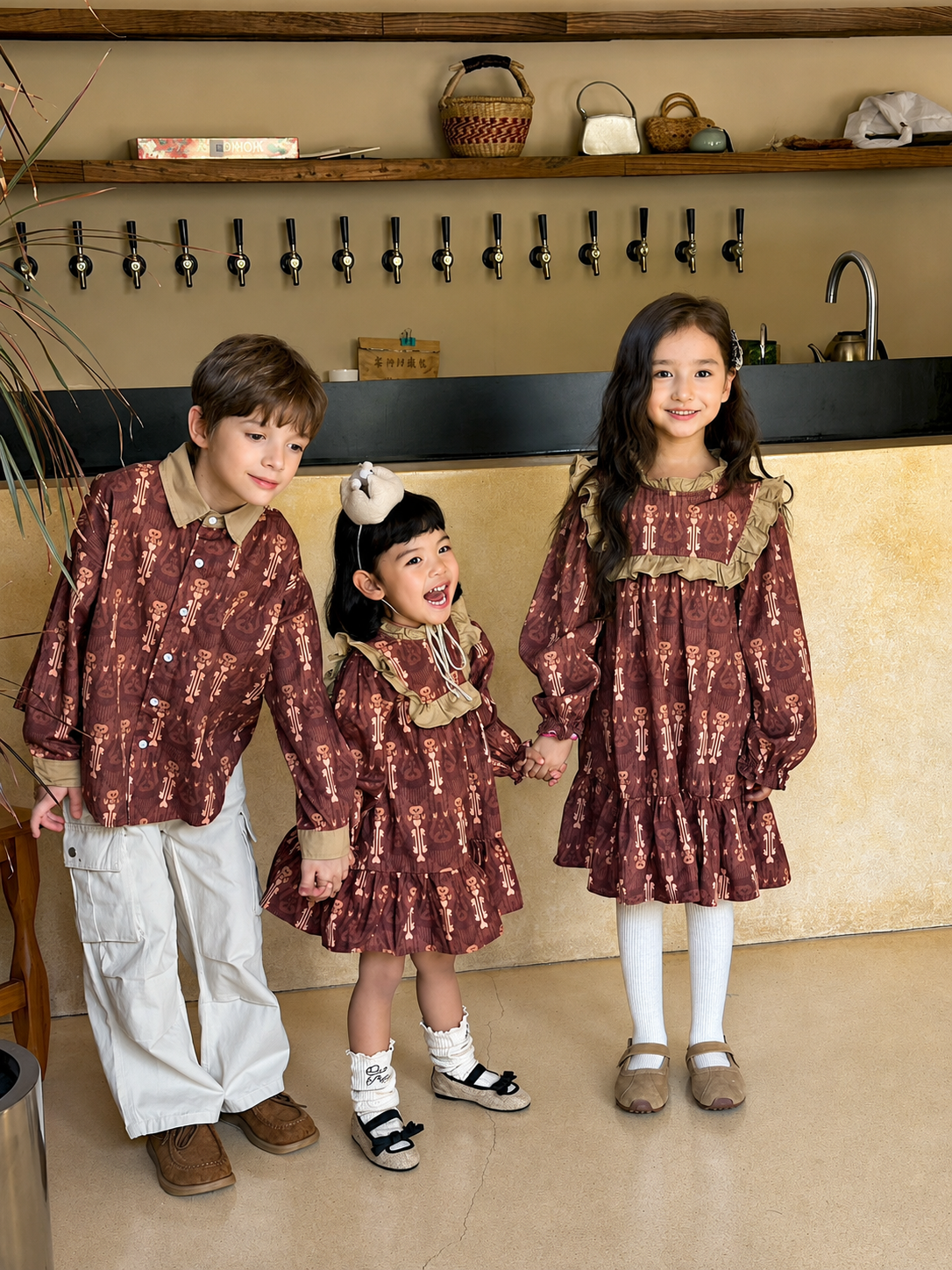 Three children in matching rust-brown cultural print outfits, a boy in a shirt and two girls in dresses, standing in a cafe.