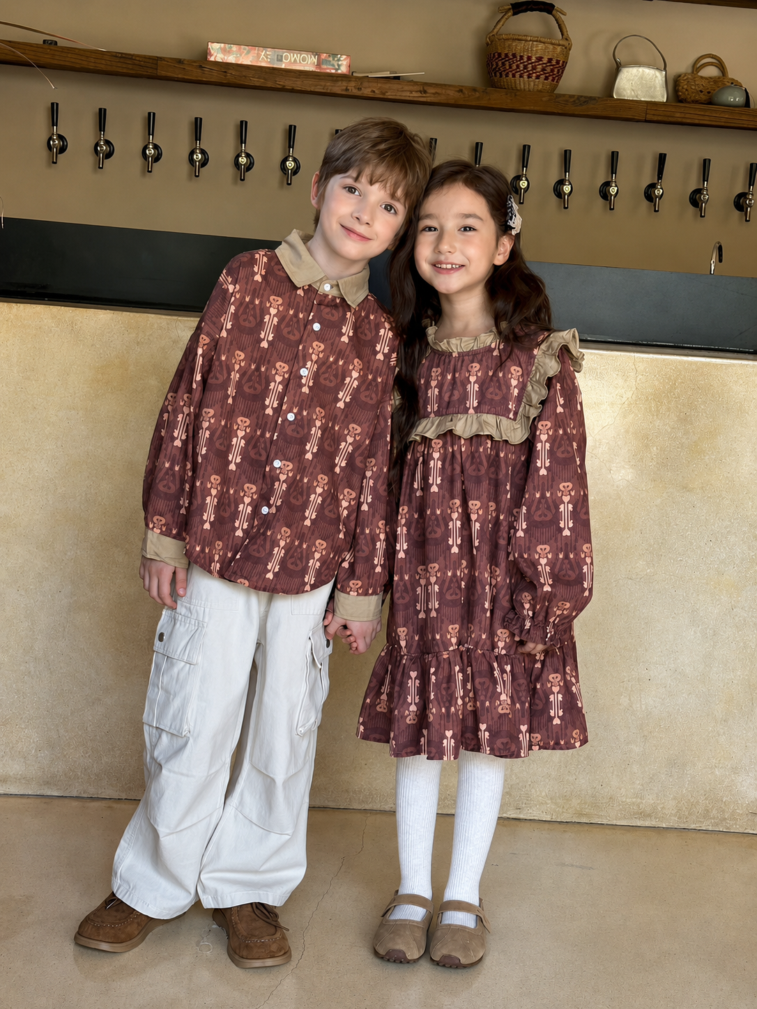 Two children wearing matching brown Atlas cotton outfits, a boy in a long-sleeve shirt and a girl in a ruffled dress, posing indoors