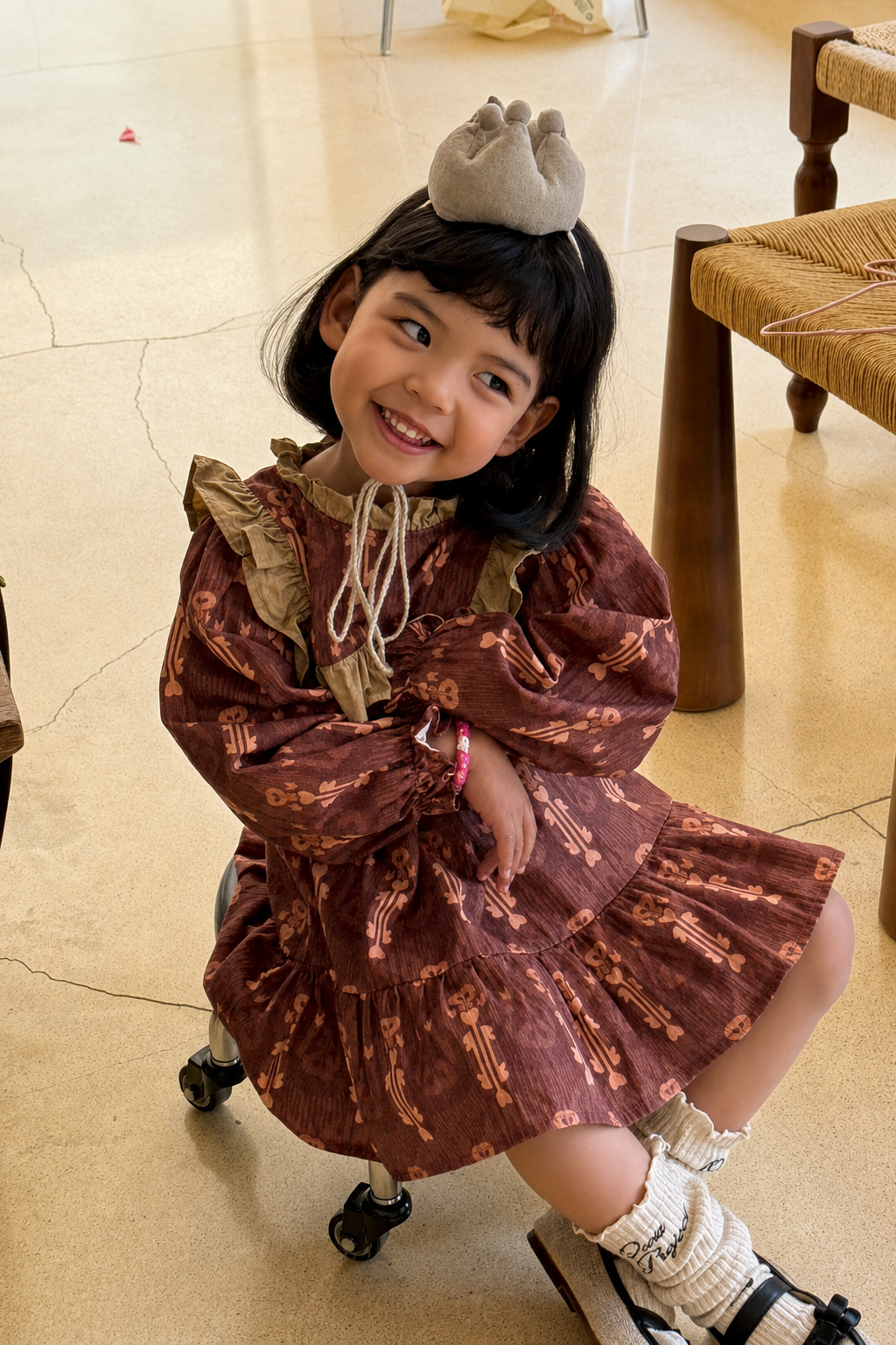 Young girl crouching and smiling in a rust-brown patterned dress with ruffled shoulders and a small crown headband.