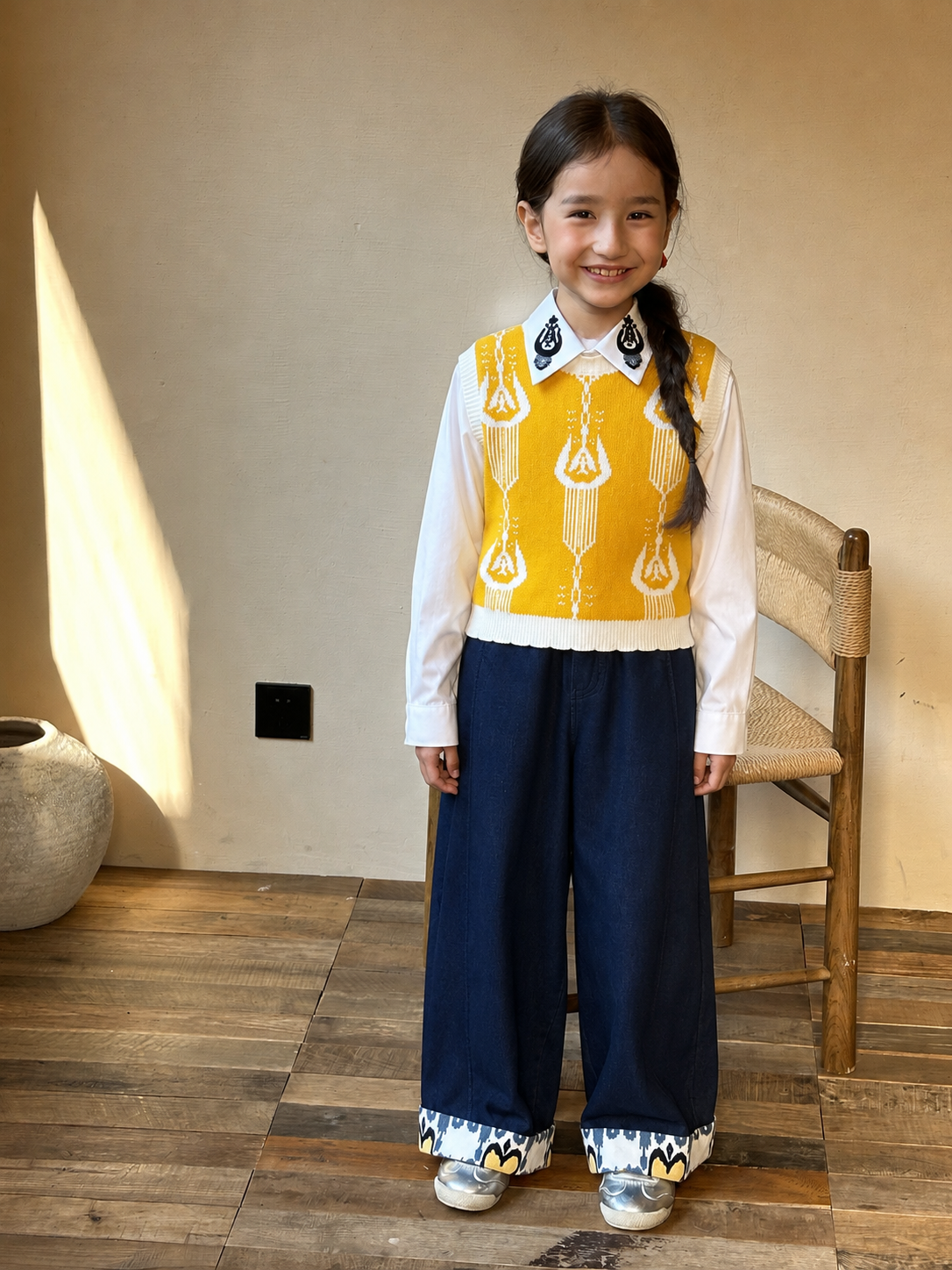 Smiling girl wearing the yellow ikat knit vest with dark navy pants standing by a chair in warm sunlight