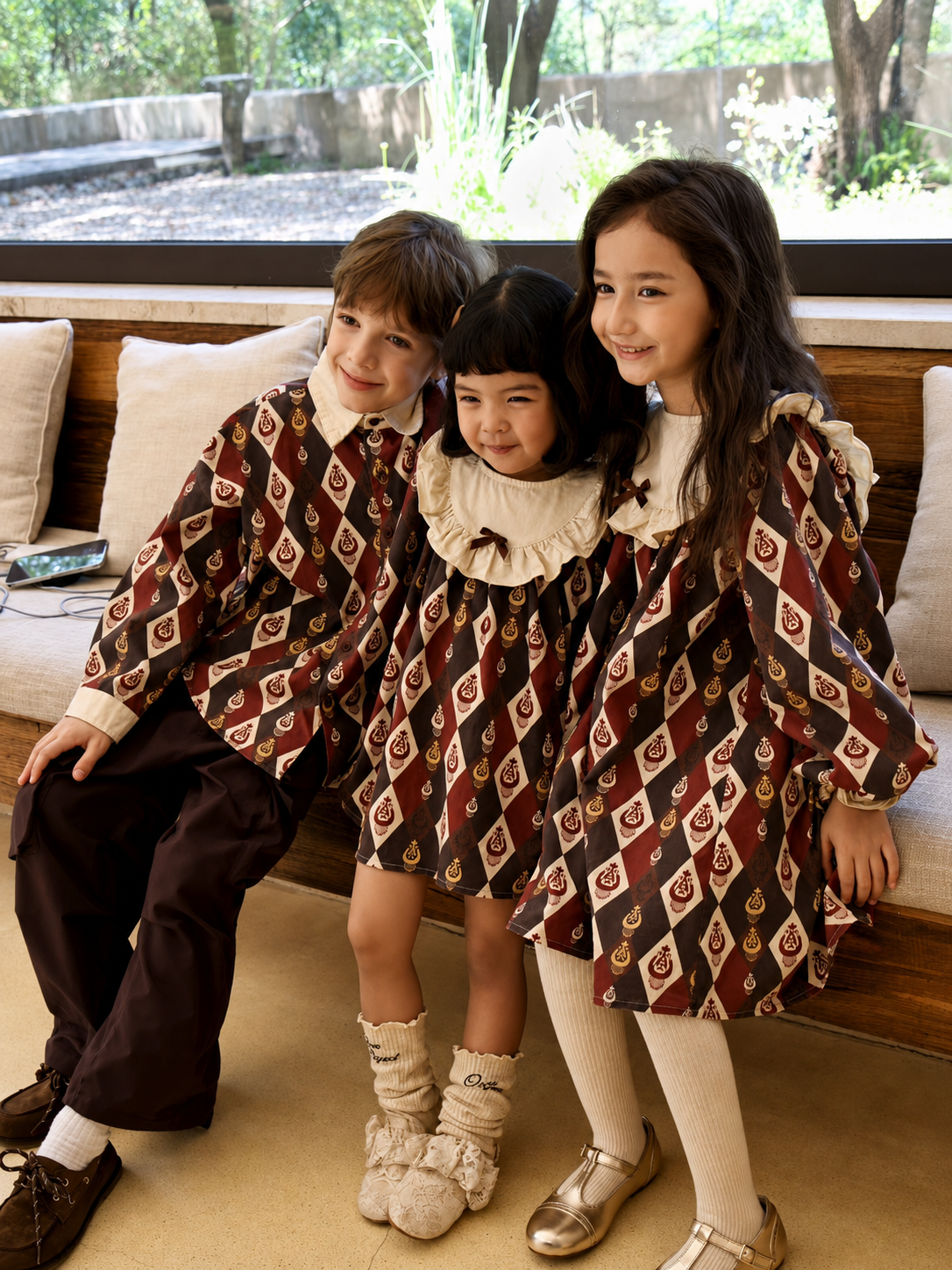 Three smiling children wearing the matching burgundy diamond-ikat outfits, sitting together on a beige sofa.