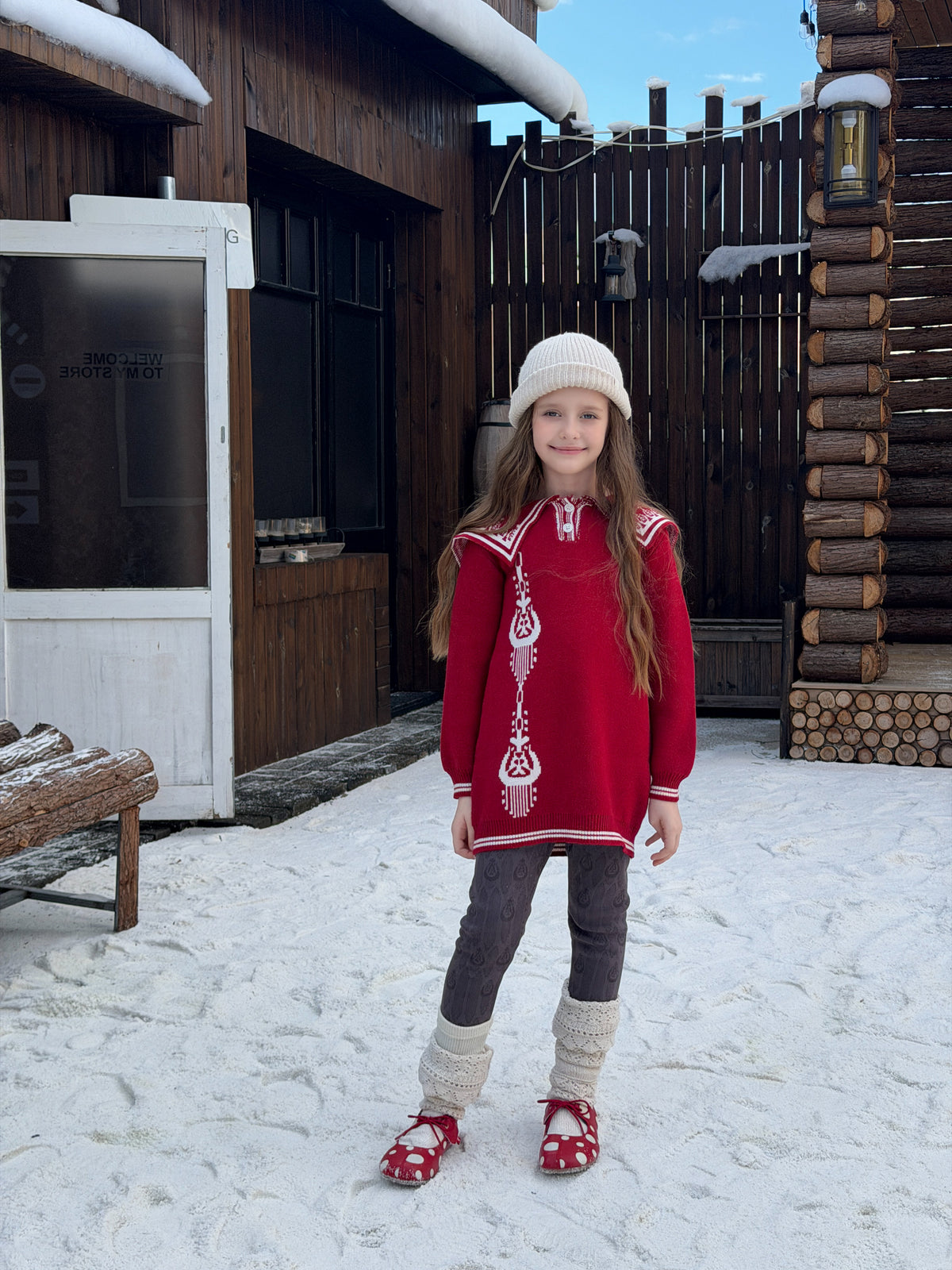 Girl wearing the red atlas knit sweater dress with white embroidered collar and a cream beanie, standing in snow by a log cabin