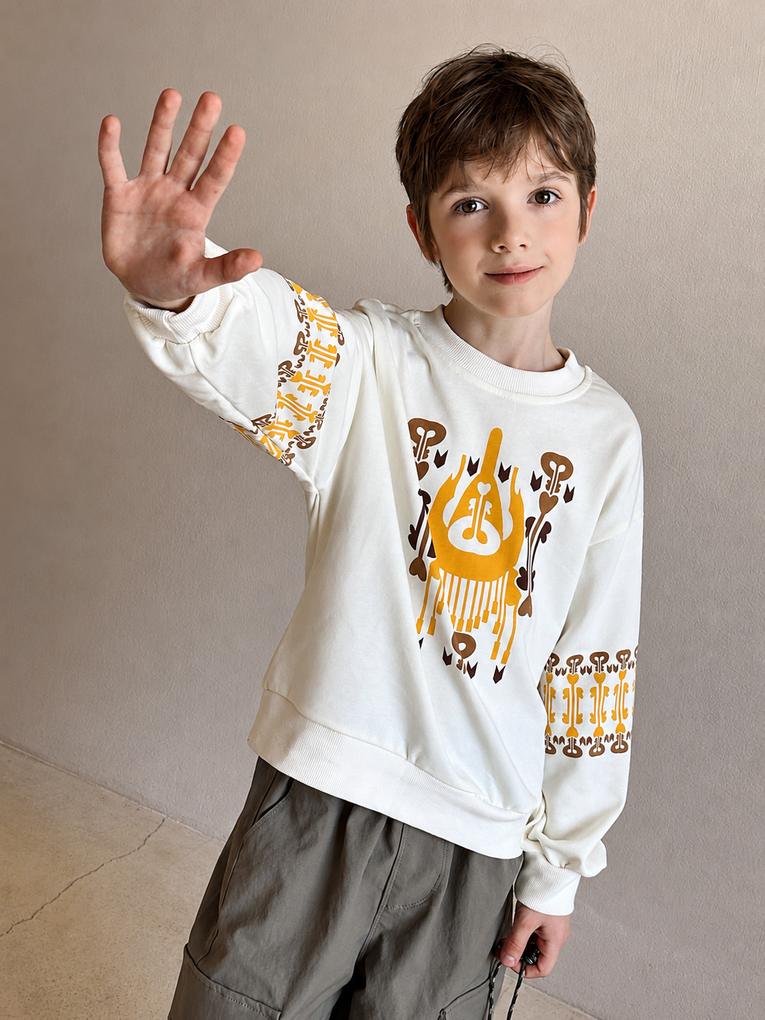 Boy waving at the camera while wearing the cream sweatshirt with orange atlas graphic and patterned cuffs indoors