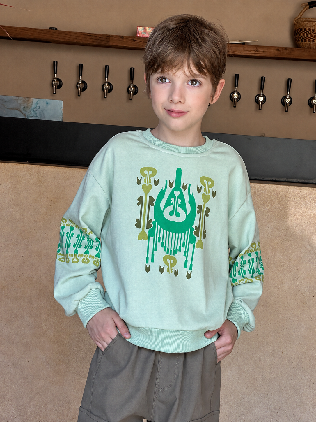Boy wearing the mint green atlas graphic sweatshirt with green ikat print, standing by a counter with taps behind