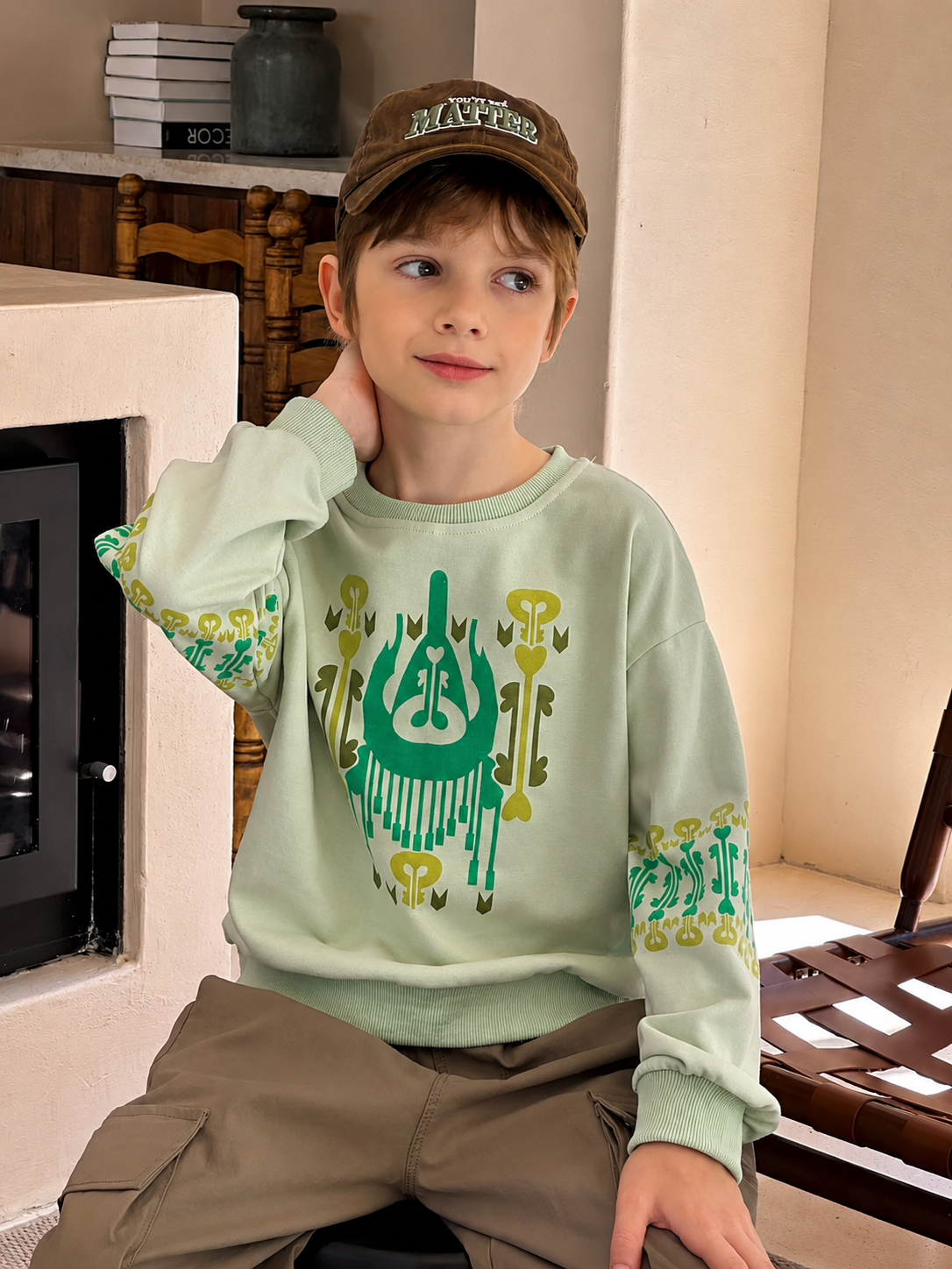 Boy in a brown cap and mint atlas graphic sweatshirt with green print, seated indoors by a fireplace
