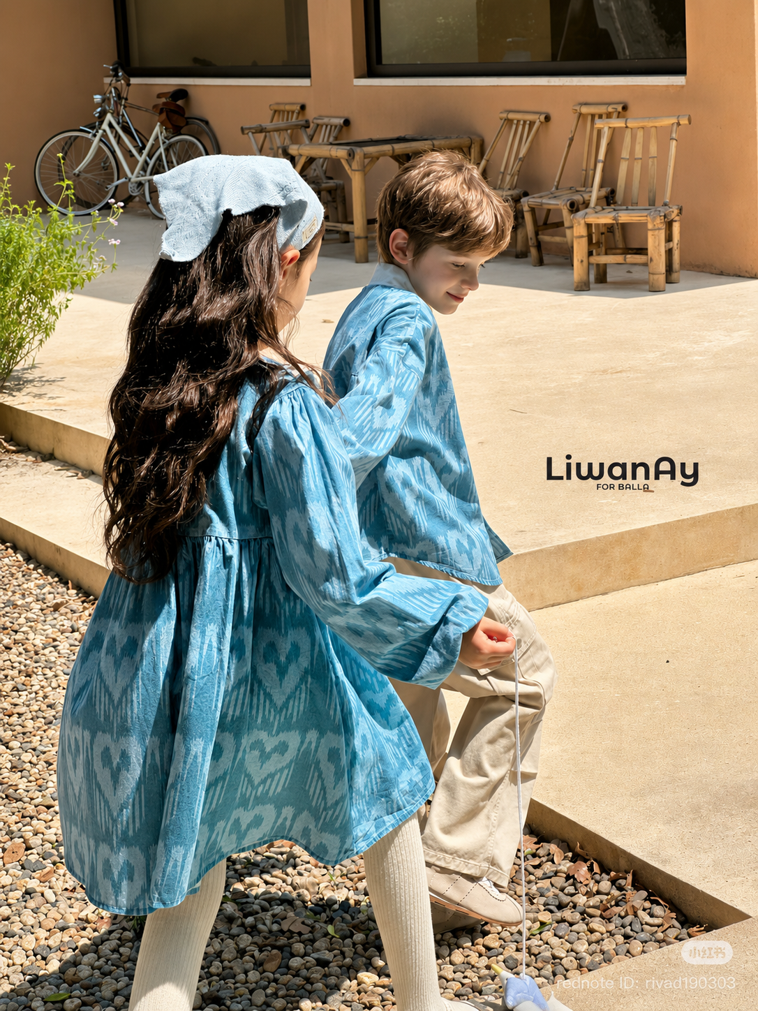 Back view of the children in blue ikat sibling outfits, the girl adjusting her headscarf near stone steps.