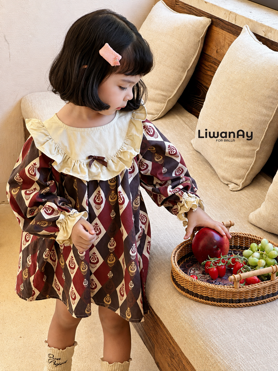 Young girl wearing the burgundy diamond-ikat dress with cream ruffle collar, standing by a sofa with a fruit basket.