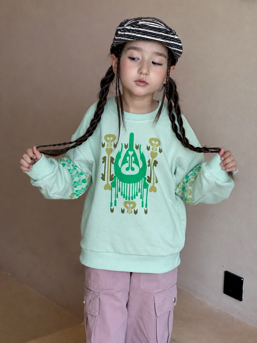 Girl with braids wearing the mint atlas graphic sweatshirt and striped cap, holding her braids indoors
