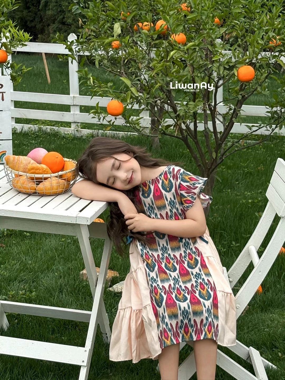 Girl wearing a colorful Liwanay summer dress outdoors beside fresh fruit and greenery.